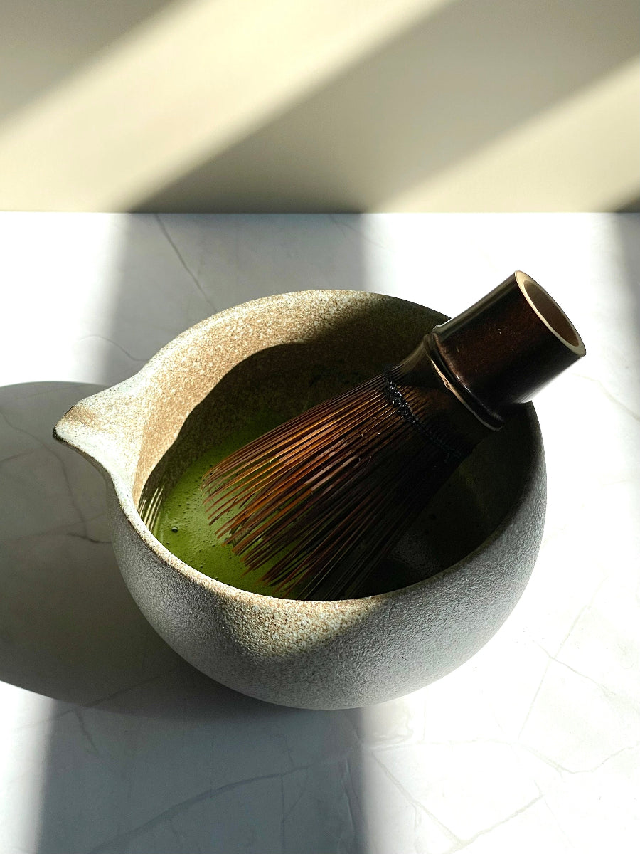 Matcha bowl with spout, filled with frothy matcha tea, with bamboo whisk inside. On marble stand.