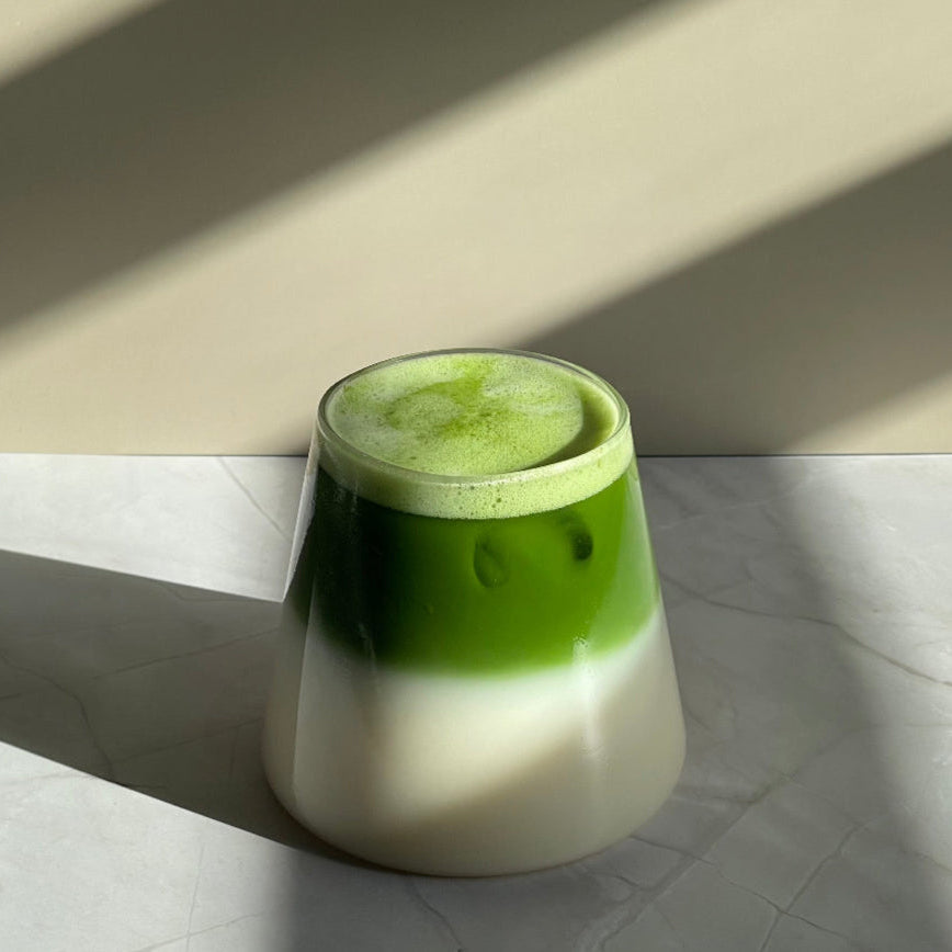 Matcha latte mousseux dans un verre conique transparent, sur supprot en marbre, avec jeu d'ombre à l'avant.