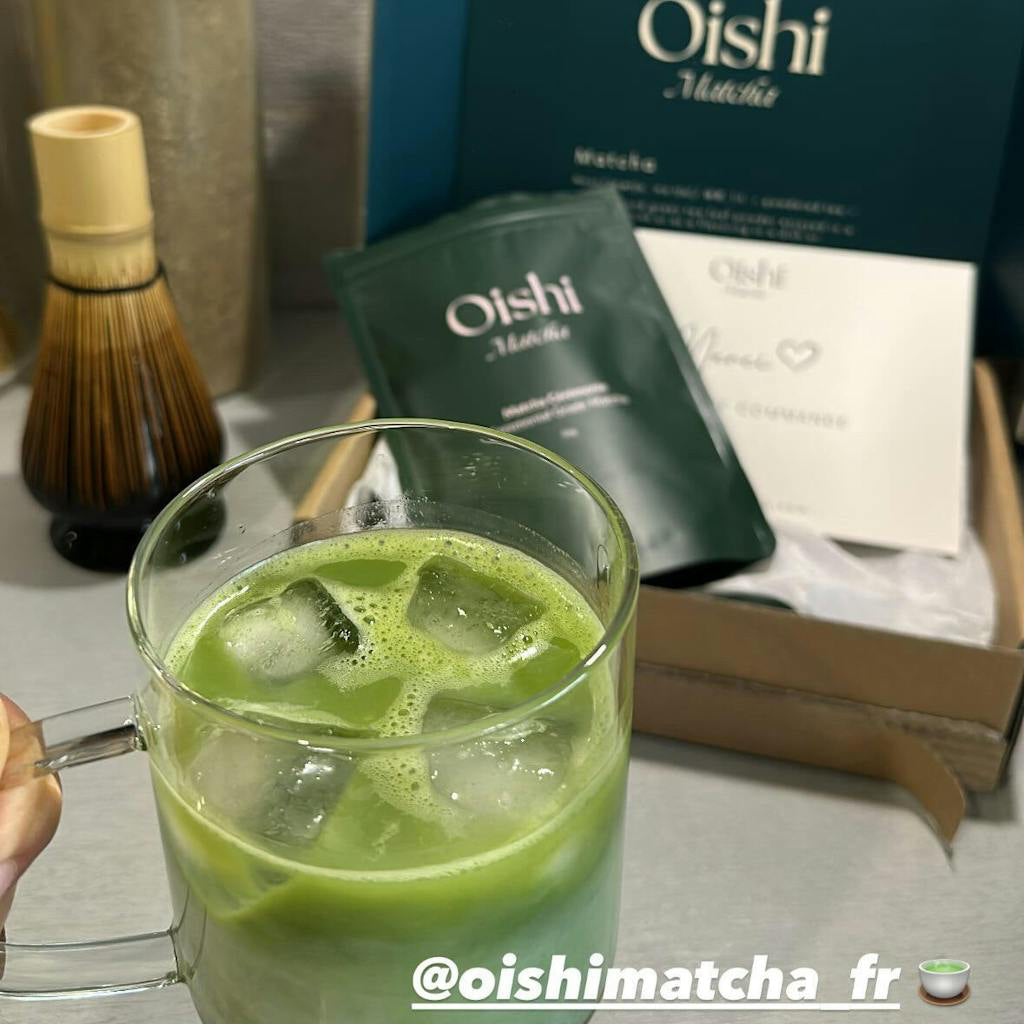 Verre de matcha latte glacé en premier plan, boite Oishi Matcha ouverte avec sachet de 50g vert et carte de remerciement, ainsi qu'un fouet en arrière plan.
