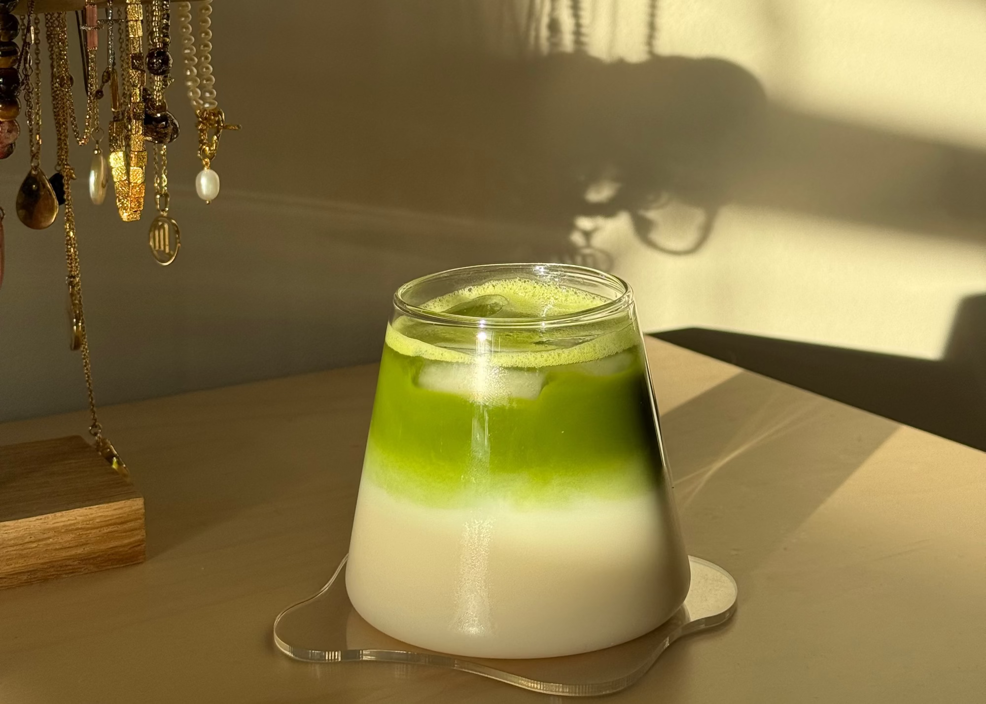 Verre de matcha latte glacé sur meuble en bois avec des bijoux en arrière plan, lumière naturelle dorée