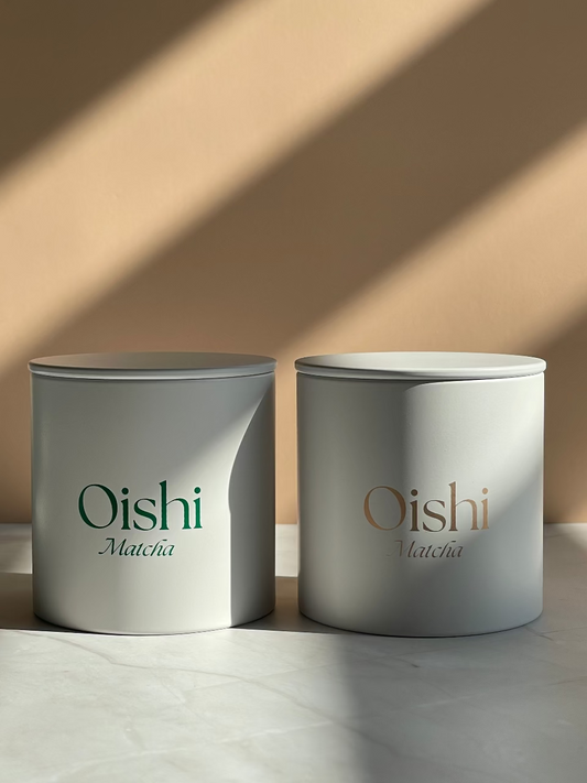 White cylindrical tea caddies, one with green Oishi Matcha logo, the other with gold Oishi Matcha logo.